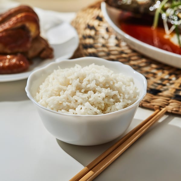 A white ceramic bowl filled with rice, accompanied by a pair of chopsticks, placed on a woven mat. Part of a meal setup.
