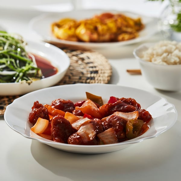 White bowl with chinese food on a table. Surrounded by dishes, utensils, and rice.