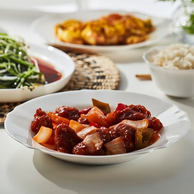White bowl with chinese food on a table. Surrounded by dishes, utensils, and rice.