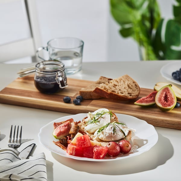 A meal on a white, wavy-edged glass plate with toast, fruit, and jam, placed on a wooden board with cutlery.