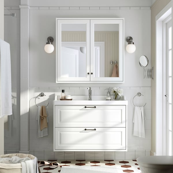 TÄNNFORSEN bathroom cabinet with mirrored doors, lights, and towels. Vase with flowers on vanity. Minimalist, bright and elegant.
