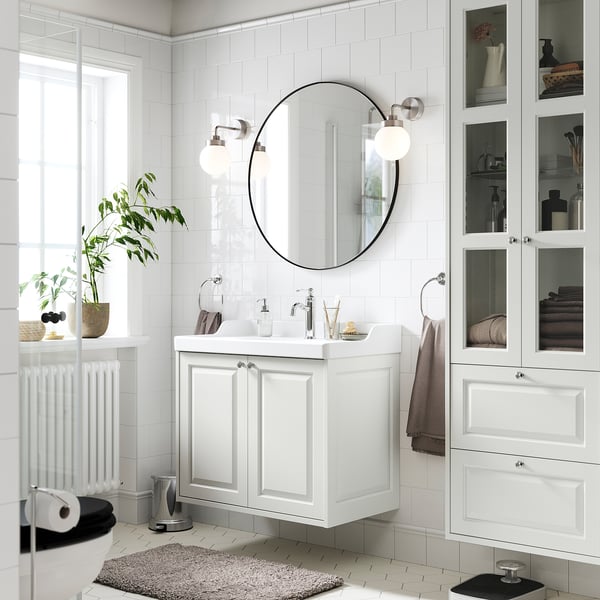 Modern white bathroom with TÄNNFORSEN vanity, round mirror, & wall lights. Plants & towels decorate, providing storage & style.