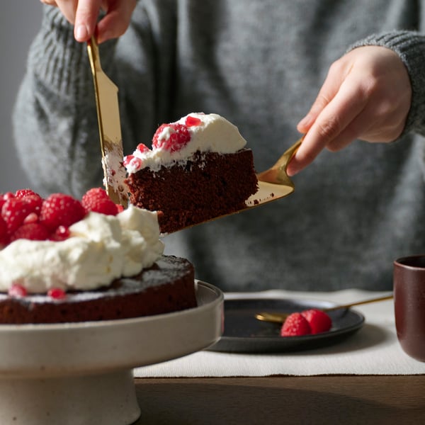 Person slicing chocolate cake with whipped cream and raspberries using a brass-colored serving spatula.