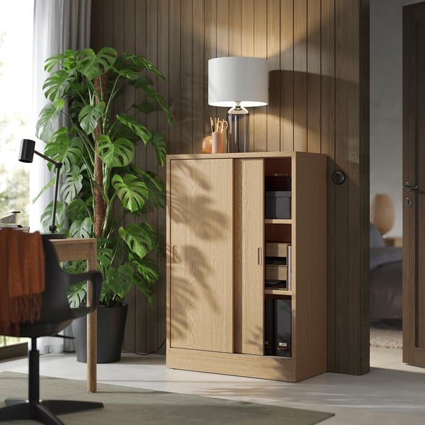 TONSTAD light oak cabinet with sliding doors and adjustable shelves. Next to a houseplant, under a lamp, in a modern room.