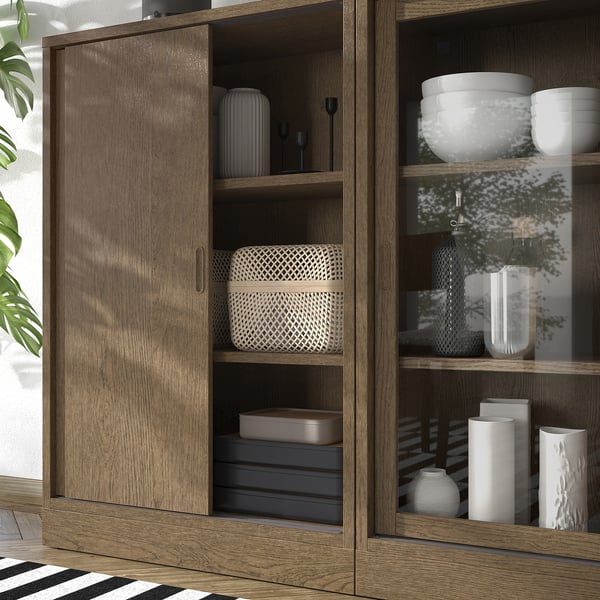 Wooden cabinet with glass door open, displaying vases, bowls, and books. Sliding door design saves space, with adjustable shelves visible.