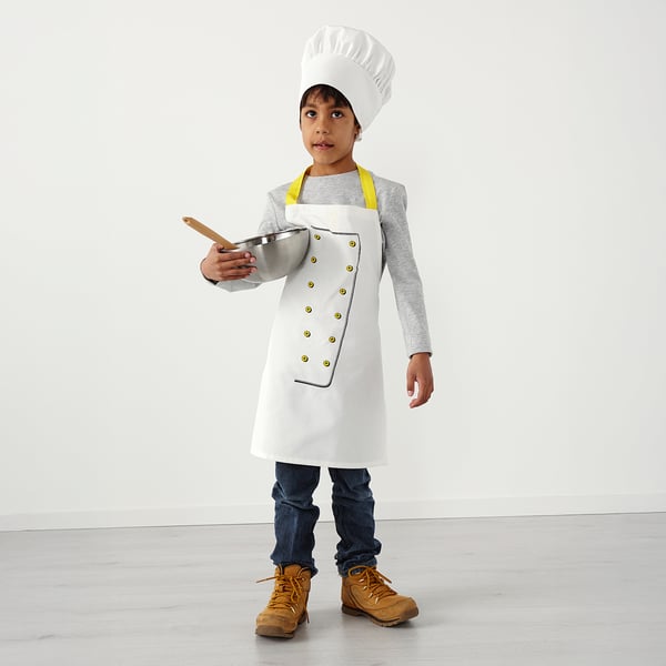 Child in chef hat & apron holding tray, role-play cooking.