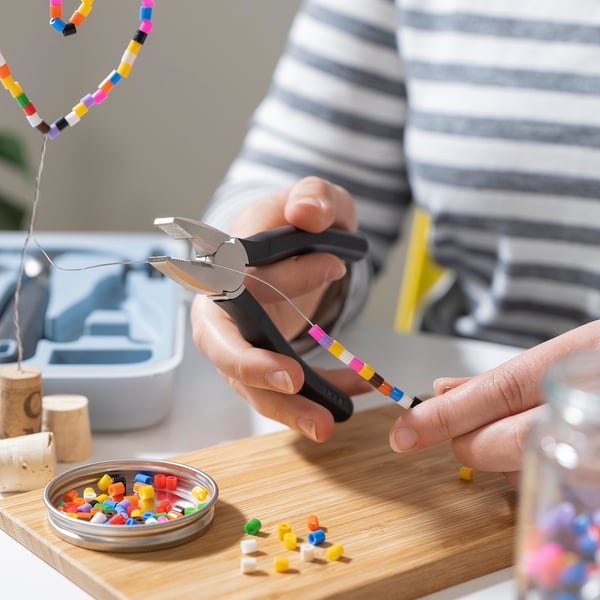 Person is using combination pliers from TRIXIG tool set to bend a colourful wire. Wooden board with various small tools and beads around.