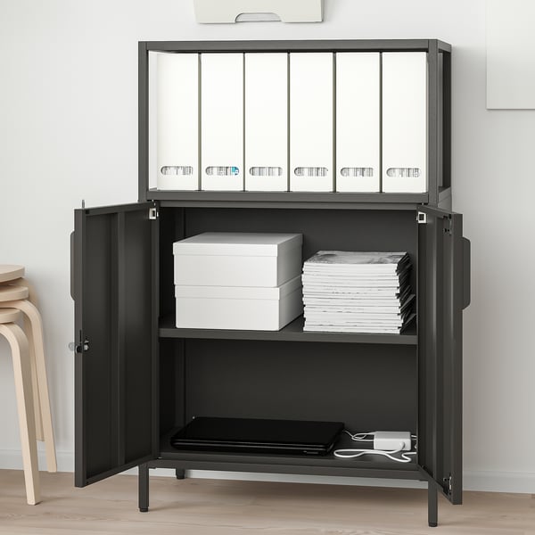 Black TROTTEN storage unit with adjustable shelves, lockable doors, and wall fastener. Holds folders, books, and a tray with chords.