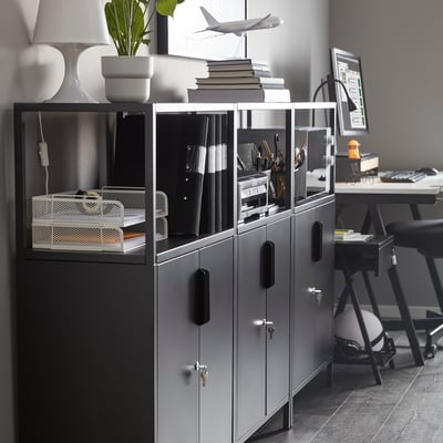 Grey TROTTEN storage unit with multiple compartments and adjustable feet, fits neatly next to a desk.