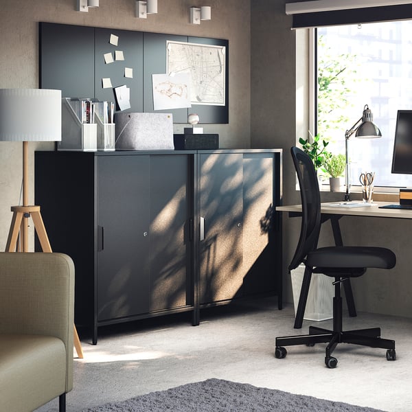 Modern office featuring TROTTEN black double-sided cabinet with sliding doors and a black office chair by a beige carpet and a large window.