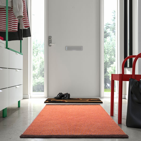 Orange VÄGOMRÅDE rug with pink/brown accents near door, under red chair, 100% cotton.