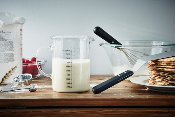 Clear glass measuring jug on wooden table with flour bag, measuring spoons, crepes, and phone.