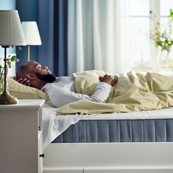 Person sleeping on a mattress with beige sheets. Beside the person is a lamp with flowers. The room has blue curtains and a window.