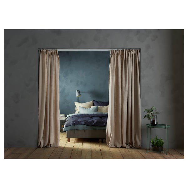 Cosy bedroom with wooden floor, blue-green walls, and light beige VIDGA curtains framing a bed with dark blue bedding. Small side table with plant.