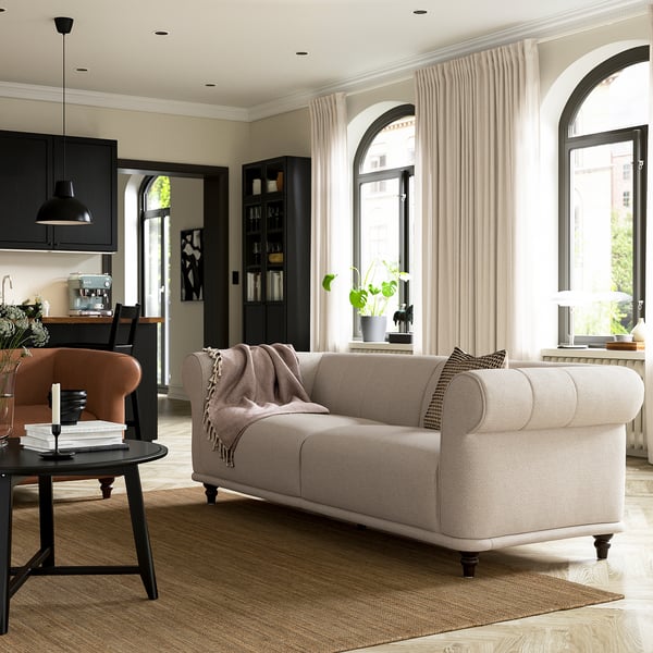 Modern living room with beige sofa, orange chair, black side table, large windows, plant, and kitchen with black cabinets and white counters.