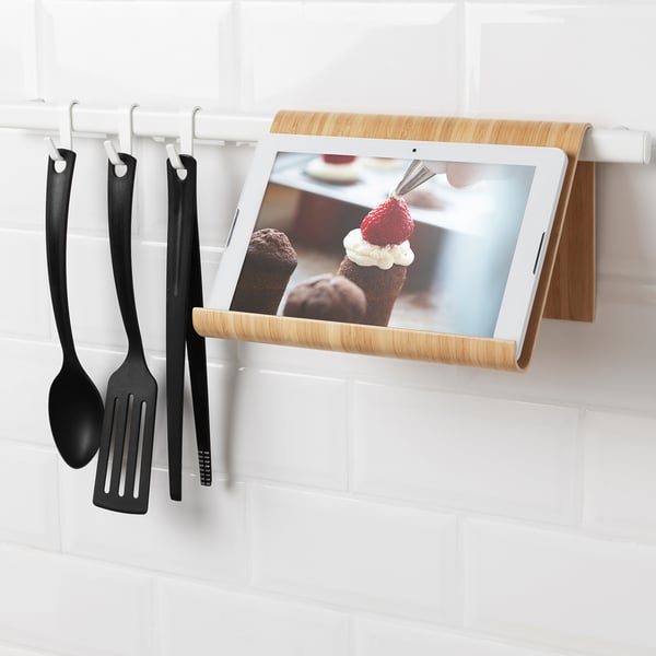 Tablet stand hangs on wall rail; holds tablet or book, freeing counter space for cooking; dark black utensils hang to the side.