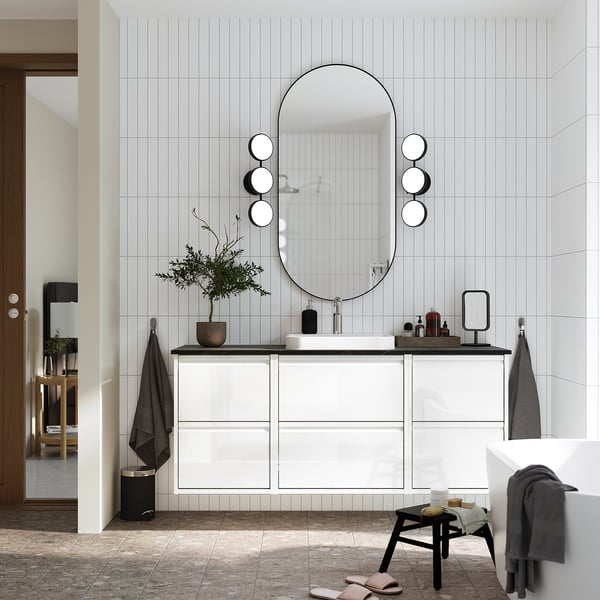 Modern bathroom with large oval mirror above white countertop unit. Three drawers, small stool, grey towels, and plant nearby.