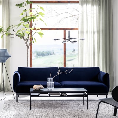 Modern living room with navy blue ÄPPLARYD sofa, glass-top coffee table, hanging light, large window, green plant, curtains, and rug.
