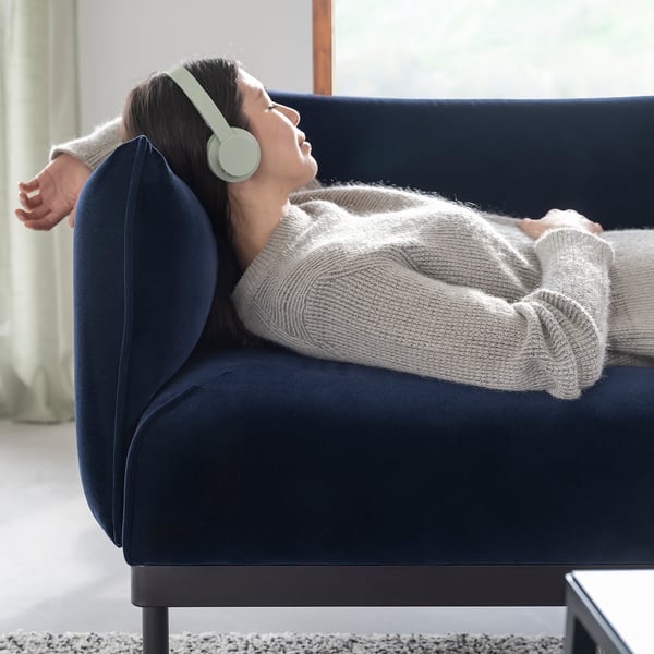 Person reclines on deep blue ÄPPLARYD sofa, wearing green headphones, cosy in grey sweater.