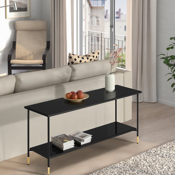 Black ÄSPERÖD coffee table with gold legs & glass top in stylish living room, holding fruit bowl, flowers, & books.