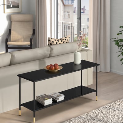 Black ÄSPERÖD coffee table with gold legs & glass top in stylish living room, holding fruit bowl, flowers, & books.