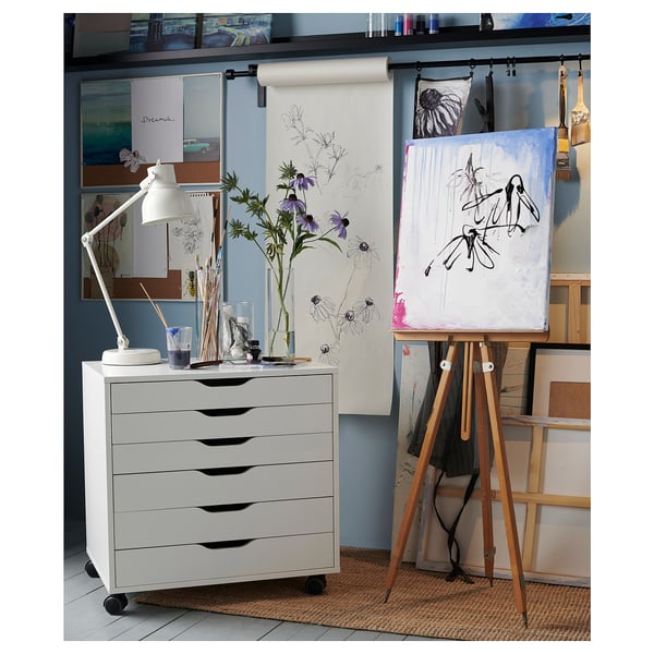 White storage unit with five drawers, wooden legs, used in an artists studio next to an easel and paintings.
