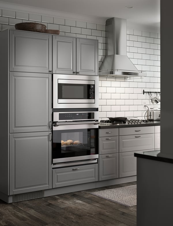 Modern kitchen with stainless steel appliances: double oven, microwave, and fridge. White subway tiles and grey cabinets. Wooden floor.
