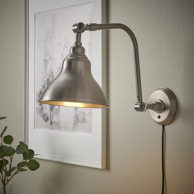 Silver adjustable wall lamp, lit and angled, with a yellow glow. Positioned next to a framed picture and a small plant.