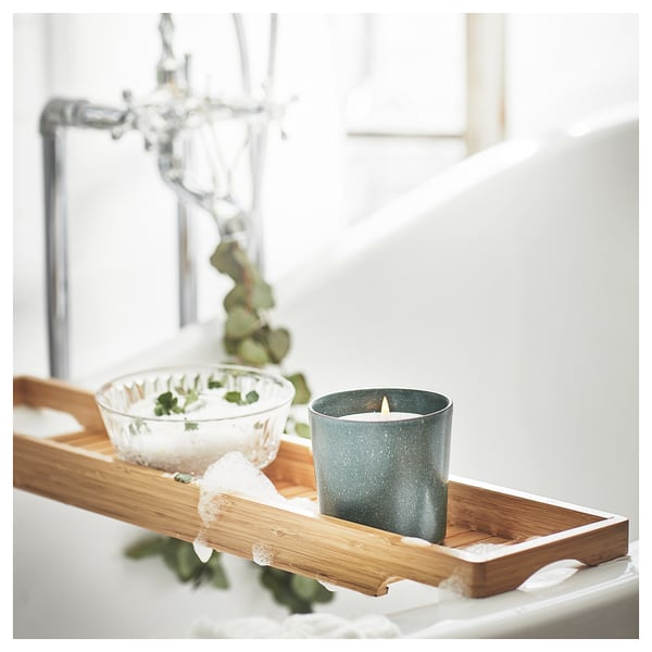 A scented green candle, AVMÅLA, sits on a wooden tray next to a glass bowl with greenery.
