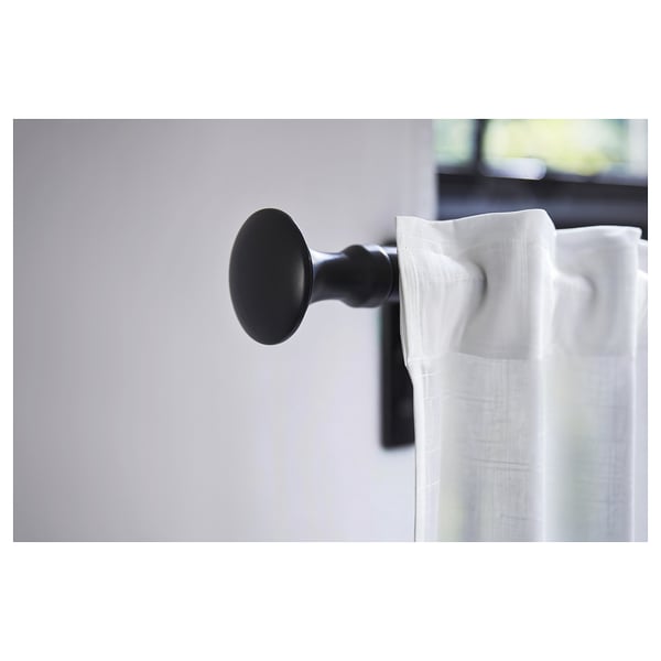 Black spherical finial attached to a white curtain rod, hanging a sheer white curtain, mounted on a light grey wall.