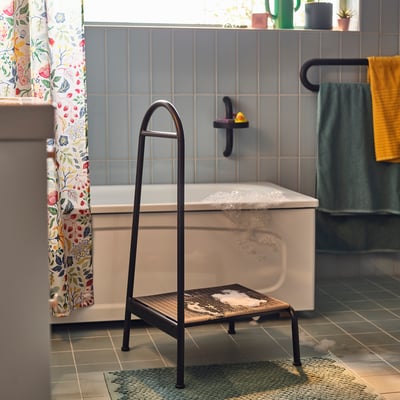 Modern grey bathroom with white bathtub, black stool, floral curtain, and yellow towel.