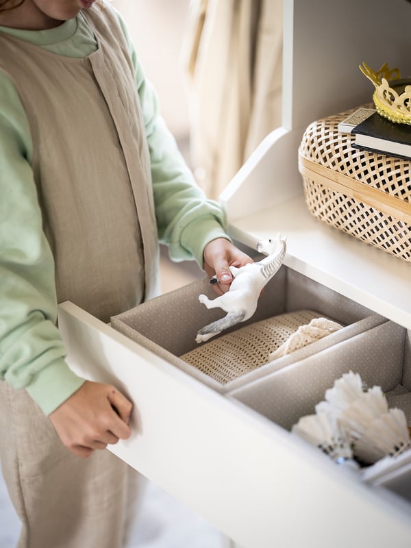 Person opens beige BAXNA fabric organiser box, revealing white items. Light green attire, indoors, near window.