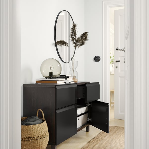 Black BESTÅ sideboard with open door and drawer, featuring mirror, globe lamp, books, vase with plant, basket with blanket in a modern room.
