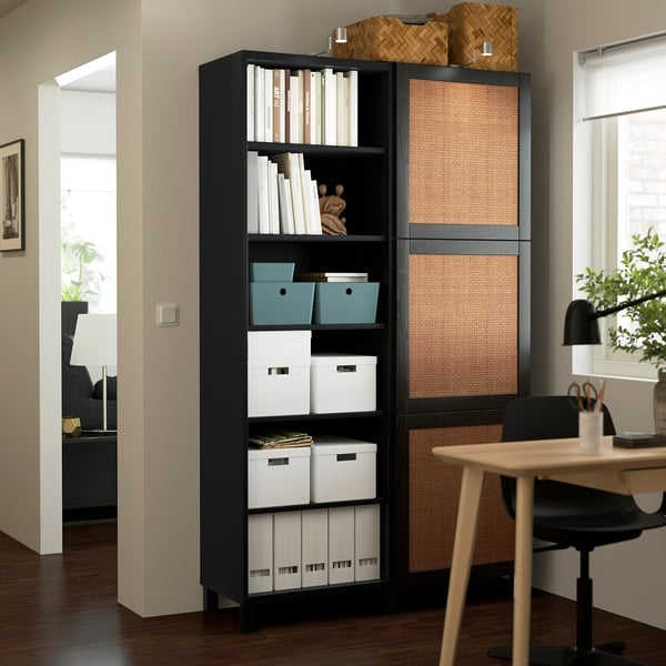 A BESTÅ black bookshelf, holding storage boxes and books. Its tall and placed between a desk and doorway, with woven baskets on top.
