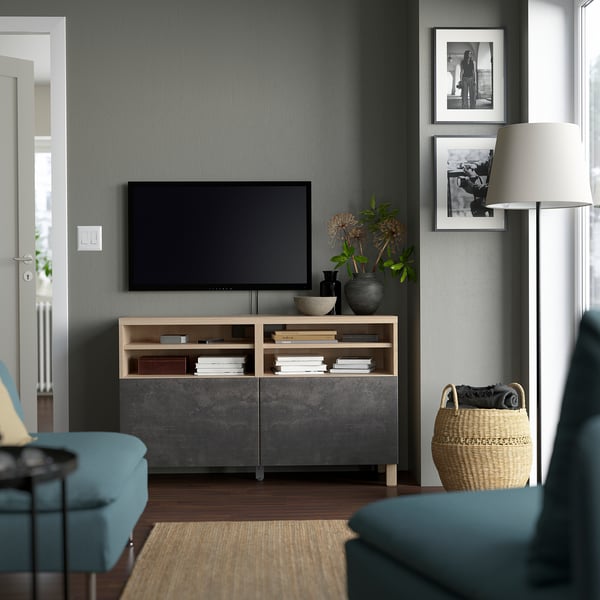 Modern tv room with black BESTÅ unit, flat-screen tv, books, and decor. Cosy setup with rug, lamp, turquoise chair, and wicker basket.