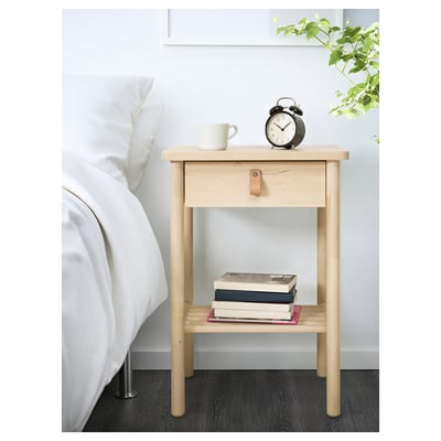 Wooden bedside table with drawer and shelf, holding books, a mug, and an alarm clock next to a white bed.