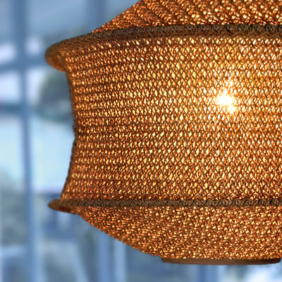 Close-up of intricate woven pendant lamp, casting warm light. Golden-hued with detailed mesh pattern.