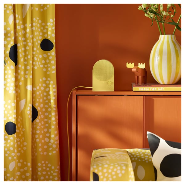 Brightly coloured curtain with yellow, white, and black dots hangs near a wooden cabinet displaying a yellow speaker and striped vase.