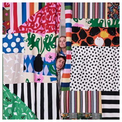 Two people stand, smiling, surrounded by colourful, patterned fabric swatches in various designs including dots, stripes, and abstract patterns.