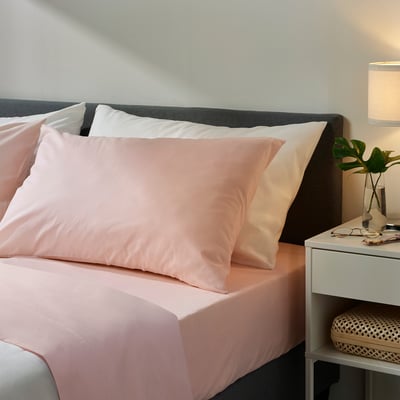 Bed with pink DVALA cotton sheets, including flat sheet and pillows, on dark headboard. Small bedside table with lamp and plant beside it.