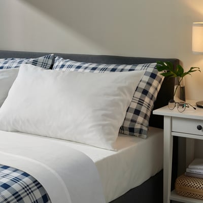 Bed with white sheets, blue plaid pillows, a side table, lamp, and plant. Headboard simple and modern.