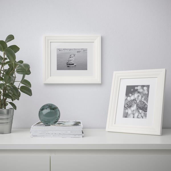 White shelf with two picture frames, a plant, and decorative items. One frame shows a sailboat, the other a butterfly.