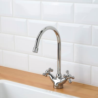 A chrome tap with cross handles and a curved spout, mounted on a wooden counter beside a sink, set against a white tiled wall.