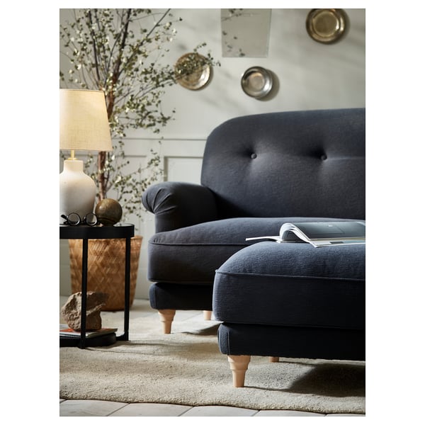 Dark blue, plush sofa with wooden legs and matching footstool, placed on a textured rug. Decorative side table with lamp and plant nearby.