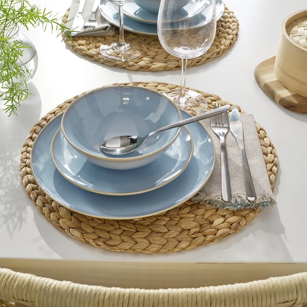 Round woven placemat with blue plates, bowl, utensils, and wine glass.