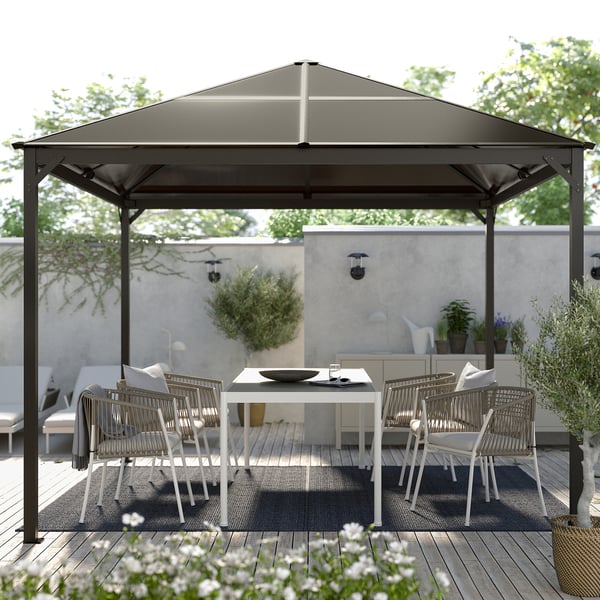Modern black gazebo with white chairs and table on a wooden deck, surrounded by greenery.