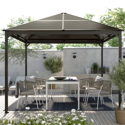 Modern black gazebo with white chairs and table on a wooden deck, surrounded by greenery.