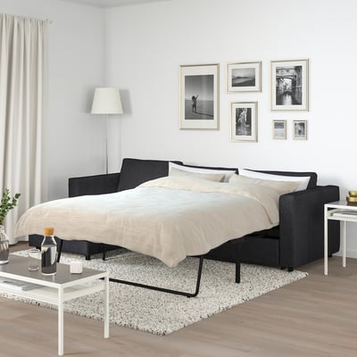 Black FINNALA sofa-bed with cool grey upholstery, under black and white photos, near white lamp.