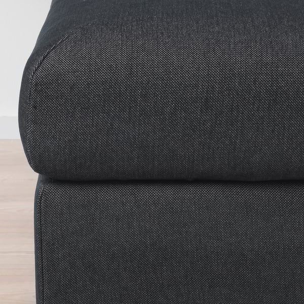 Black fabric footstool with plush, textured upholstery and a muted sheen, positioned on a light wooden floor.