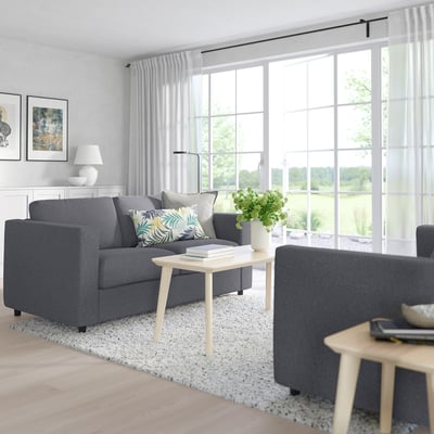 Modern living room: FINNALA grey sofa-bed, tropical pillows, wooden coffee table, white lamp, green view.
